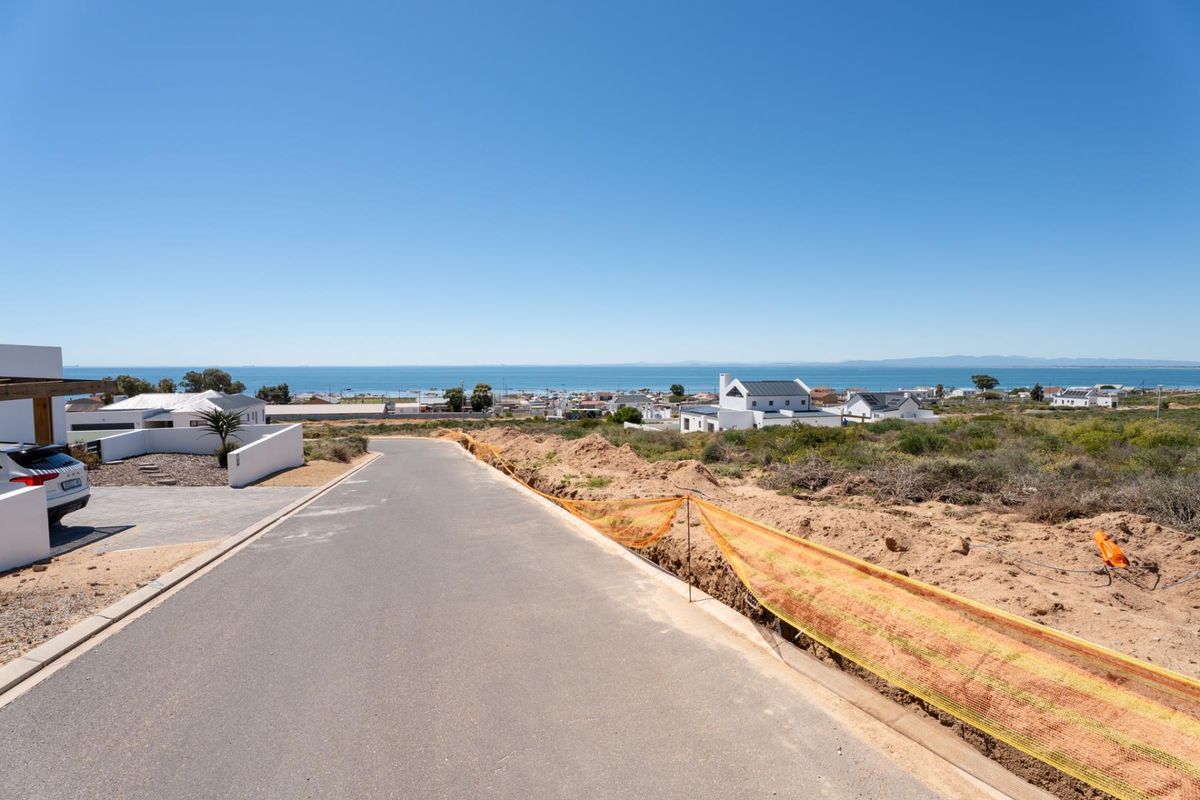 A street-level view of a paved road in a residential area, featuring clear coastal views and adjacent undeveloped land with temporary construction fencing.