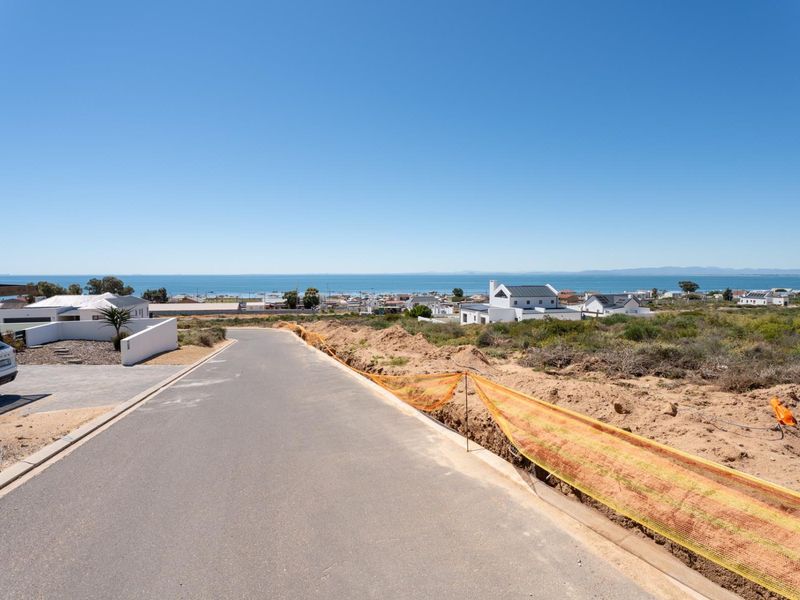 A street-level view of a paved road in a residential area, featuring clear coastal views and adjacent undeveloped land with temporary construction fencing.