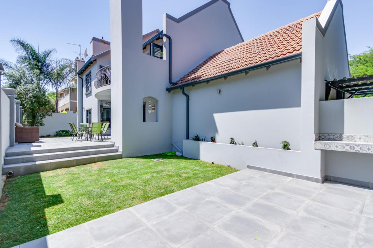 A well-maintained rear exterior featuring a tiled patio, a built-in braai area, a small lawn, and a balcony overlooking the garden space.