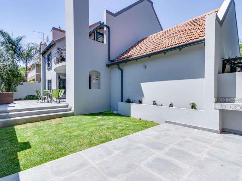 A well-maintained rear exterior featuring a tiled patio, a built-in braai area, a small lawn, and a balcony overlooking the garden space.