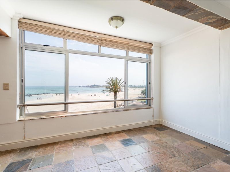 An empty living area featuring large windows with direct beach and ocean views, finished with natural slate tile flooring and a decorative ceiling beam.