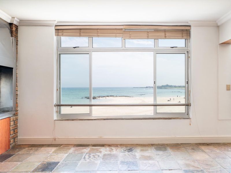 Spacious living area featuring a built-in braai, stone tile flooring, and large windows offering direct ocean and beach views.