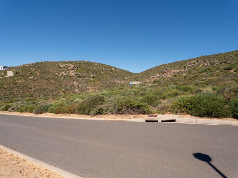 A view of the paved street frontage and concrete drainage infrastructure bordering the undeveloped residential land with natural hillside terrain.