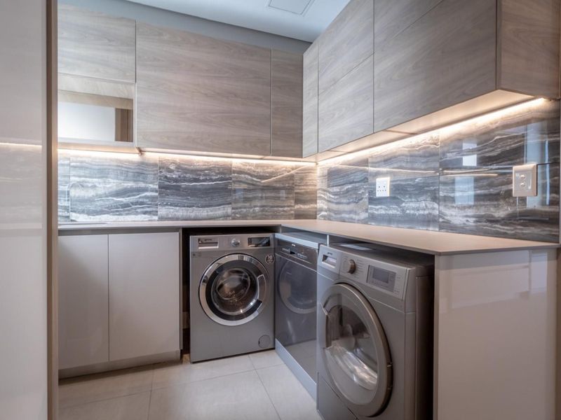 Modern, well-appointed laundry room featuring sleek wood-grain cabinetry, stone-look backsplash, under-cabinet LED lighting, and integrated sink with tiled flooring.