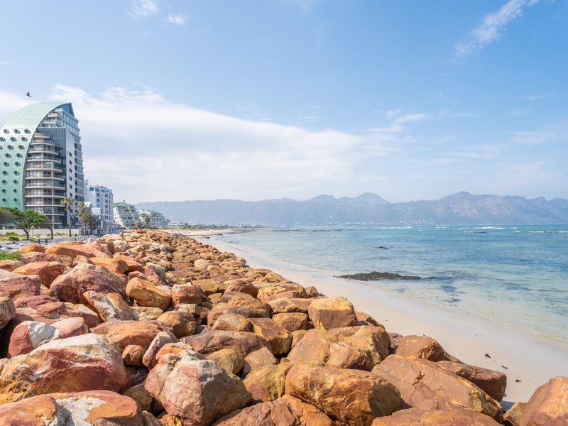 Scenic coastal view featuring a rocky shoreline, sandy beach, and distant mountain range with modern apartment buildings in the background.