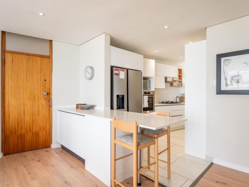Modern open-plan kitchen featuring sleek white cabinetry, stone countertops, a breakfast bar, and integrated appliances, seamlessly connecting to the living area.