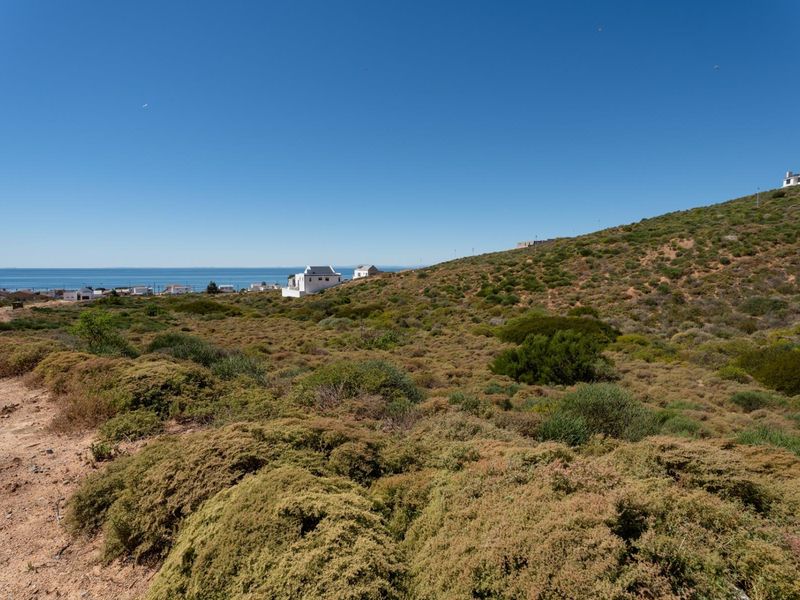 A wide-angle view of a sloped, undeveloped residential plot featuring natural coastal vegetation and distant ocean views.