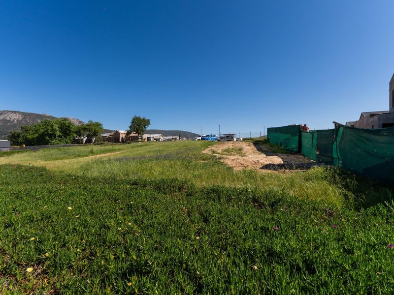 A vacant residential plot featuring natural ground cover and temporary construction fencing, situated in a developing neighborhood with mountain views.