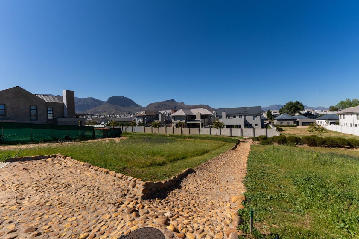 A vacant residential plot featuring a stone-paved pathway, established grass, and perimeter boundary walls with mountain views in a residential estate setting.