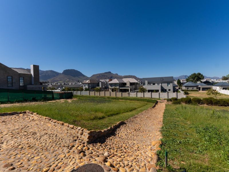 A vacant residential plot featuring a stone-paved pathway, established grass, and perimeter boundary walls with mountain views in a residential estate setting.