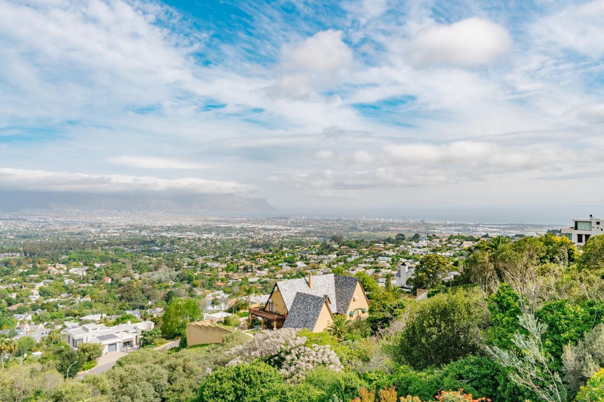 An elevated aerial view showcasing the property's hillside location, featuring a pitched roof house with a balcony overlooking the surrounding suburban landscape and distant mountains.