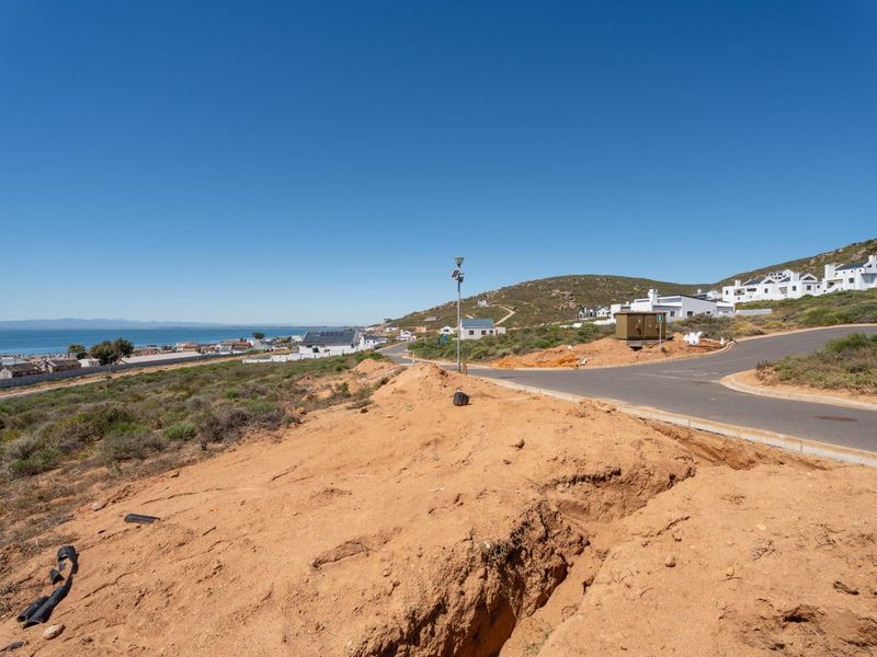 A view of a vacant residential plot featuring an excavation trench, adjacent paved road, and surrounding coastal landscape.