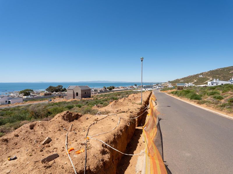 A street-level view of a residential development site featuring an asphalt road, utility trenching, and coastal views in the background.