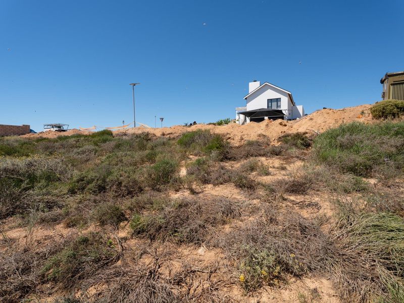 A view of a vacant residential plot featuring sandy terrain and native coastal vegetation, with neighboring residential development visible in the background.