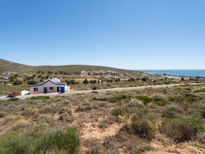 An elevated view of a vacant residential plot featuring coastal landscape, nearby road access, and surrounding neighborhood scenery.