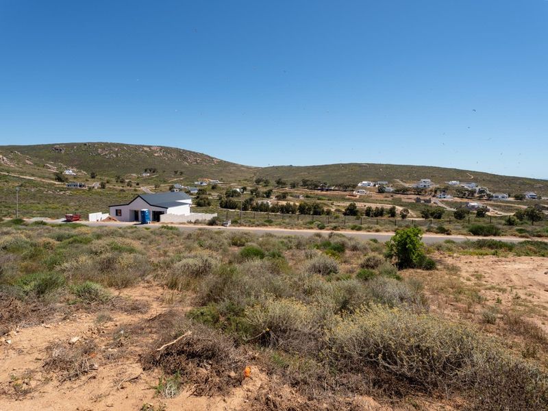 An elevated view of a vacant residential plot featuring natural vegetation, adjacent paved road access, and surrounding coastal landscape.