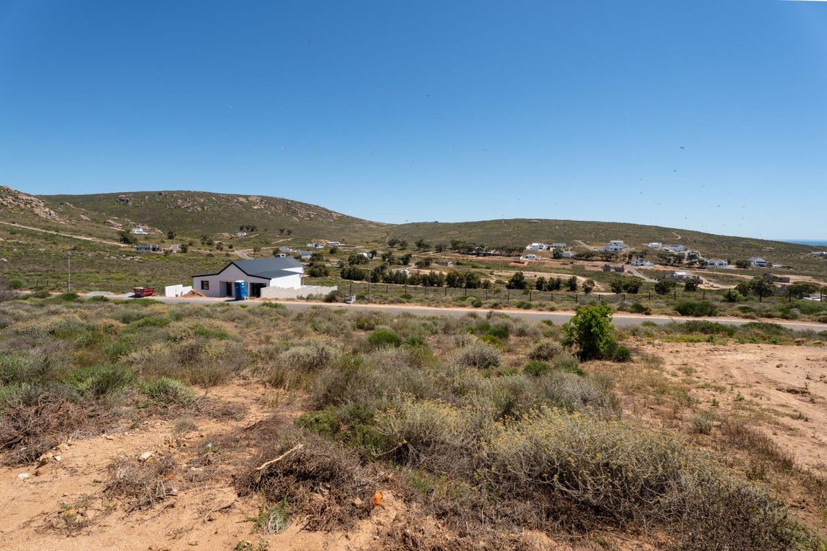 An elevated view of a vacant residential plot featuring natural vegetation, adjacent paved road access, and surrounding coastal landscape.