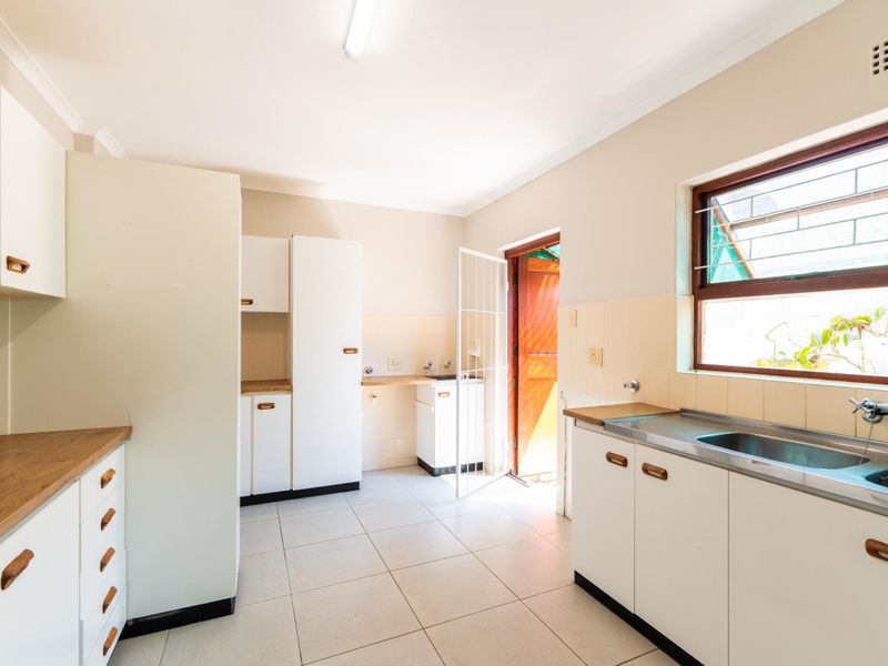 A functional, empty kitchen featuring white built-in cabinetry, laminate countertops, a stainless steel sink, and tiled flooring with direct access to the exterior.