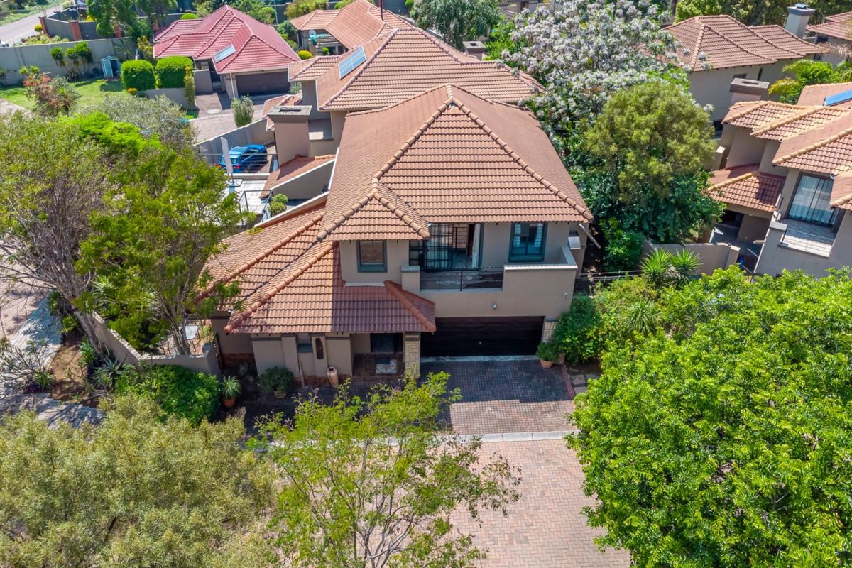 Aerial view of a residential property featuring a tiled roof, paved driveway, integrated garage, and an upper-level balcony surrounded by mature trees.