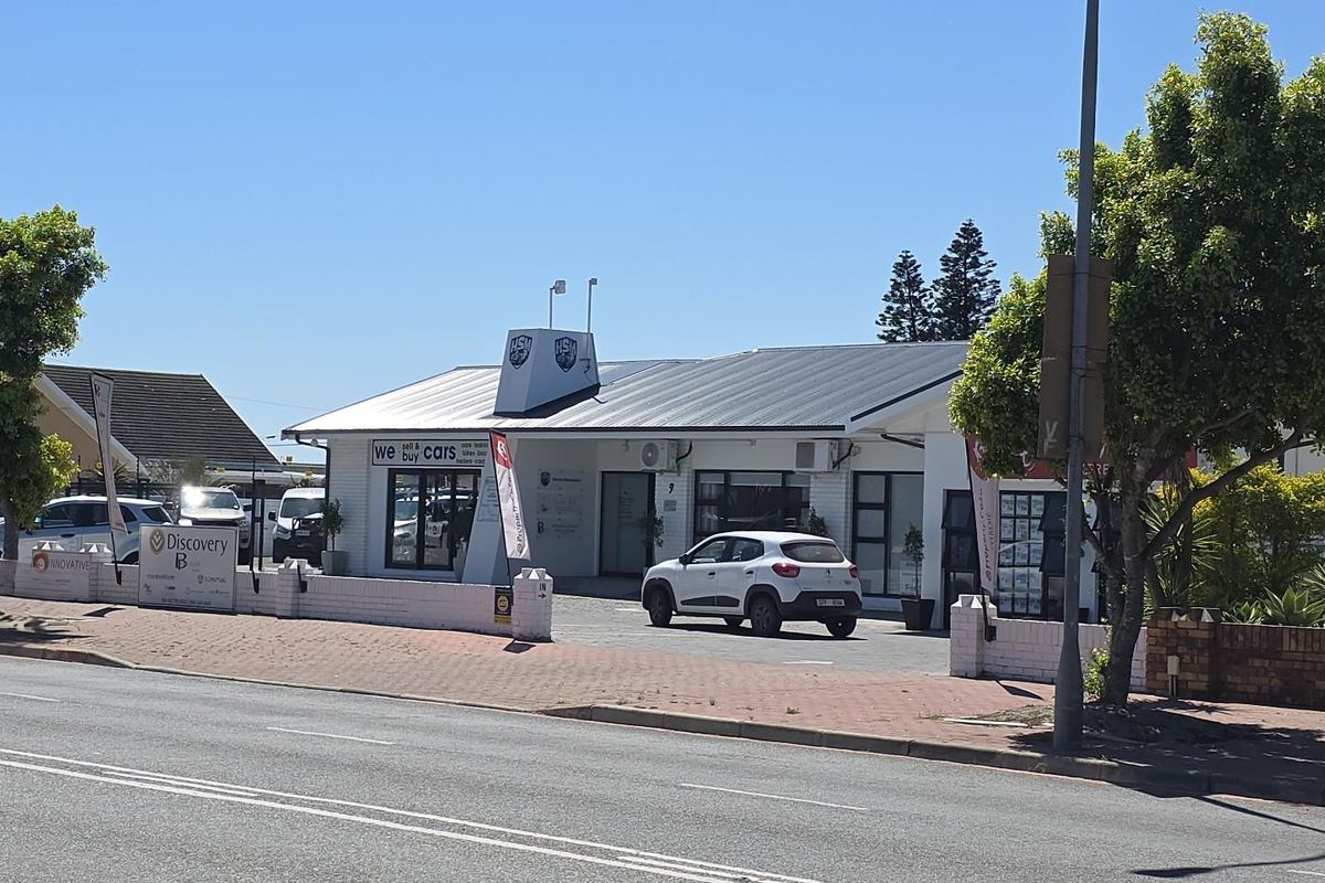 Front exterior view of a commercial property featuring a metal roof, paved driveway, and brick boundary wall, situated along a main road.