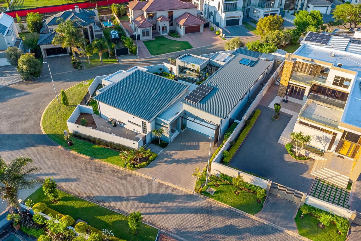 Aerial view of a modern residential property featuring a metal roof with solar panels, a paved driveway, and secure boundary walls in a suburban estate.