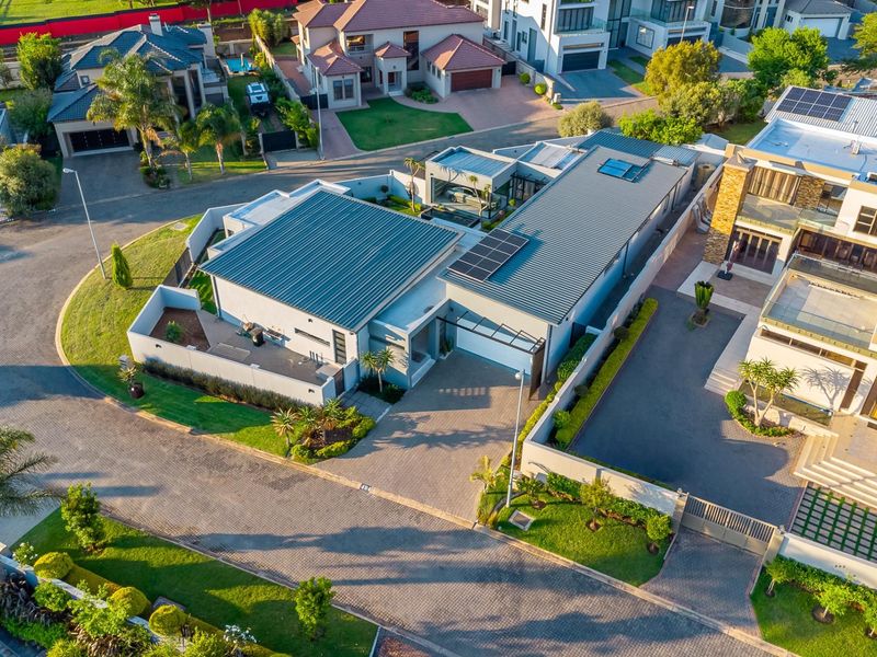 Aerial view of a modern residential property featuring a metal roof with solar panels, a paved driveway, and secure boundary walls in a suburban estate.