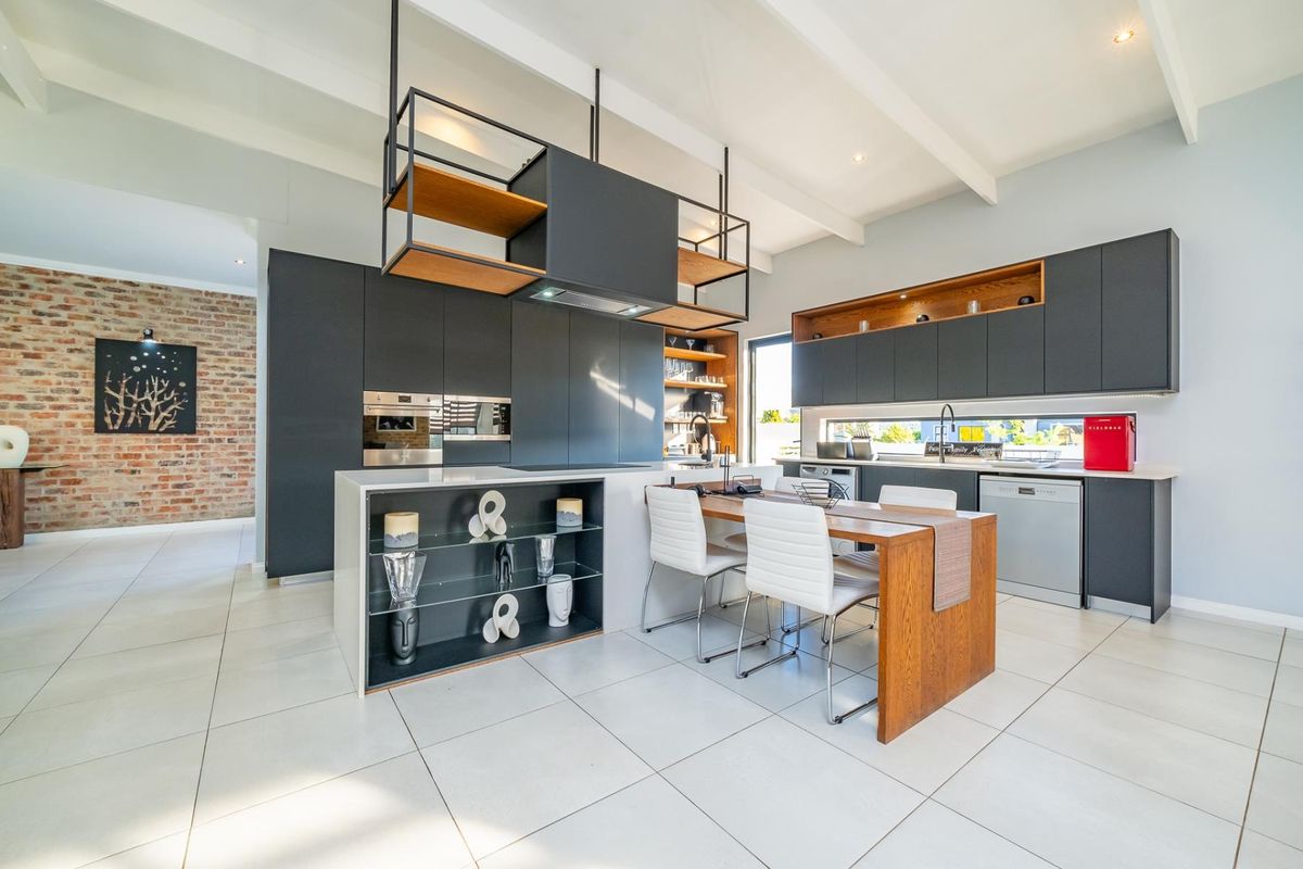 Modern kitchen featuring dark cabinetry, a central island with integrated shelving, exposed brick feature wall, and high-quality tiled flooring under exposed ceiling beams.