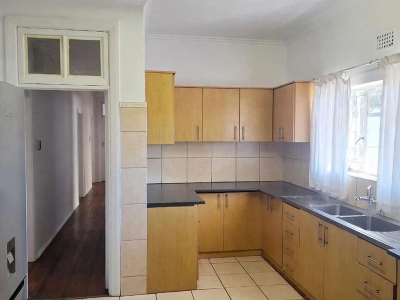 A functional kitchen featuring laminate cabinetry, dark countertops, double stainless steel sink, and ceramic tiled flooring with direct access to the hallway.