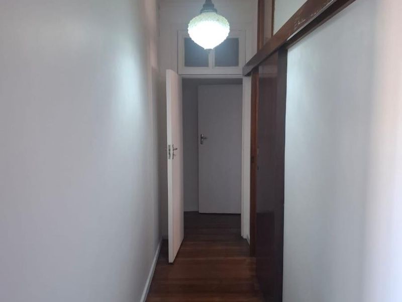 A narrow hallway featuring polished hardwood flooring, a classic pendant light fixture, and a transom window above the doorway.