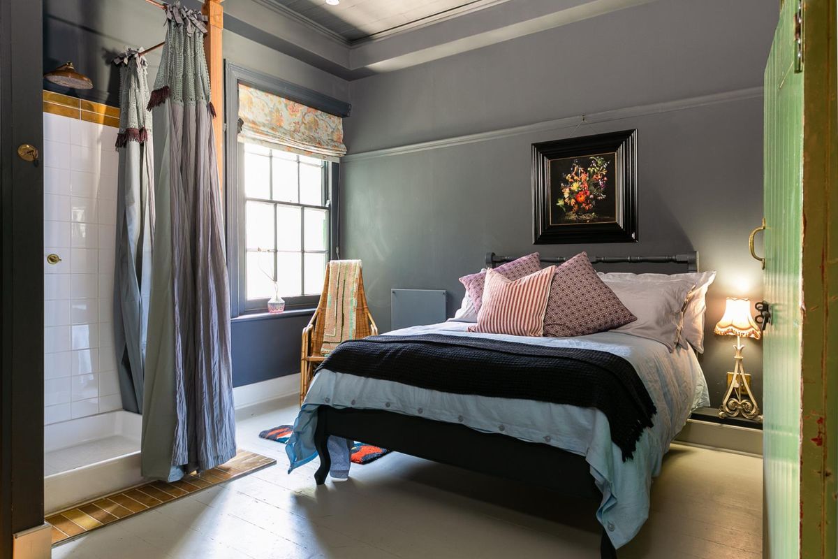 A well-appointed hotel bedroom featuring a unique integrated shower enclosure, timber flooring, and classic sash window with dark grey wall finishes.