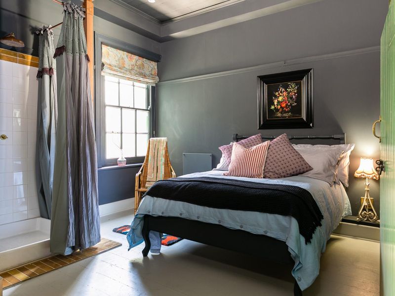 A well-appointed hotel bedroom featuring a unique integrated shower enclosure, timber flooring, and classic sash window with dark grey wall finishes.