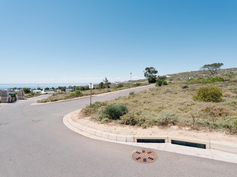 A view of the paved street infrastructure, including curbing, drainage, and a manhole cover, bordering the vacant residential land with distant coastal views.