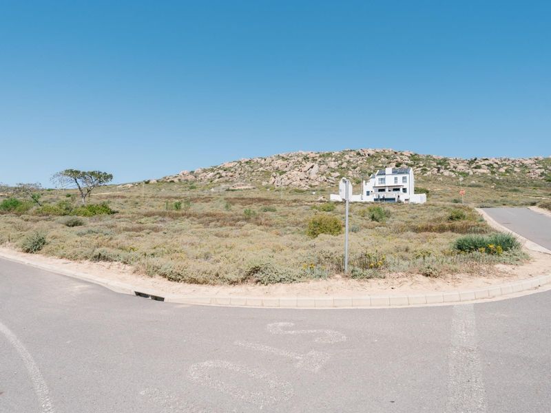 A view of the vacant residential land from the adjacent paved street, featuring established curbing and drainage infrastructure in a coastal landscape setting.
