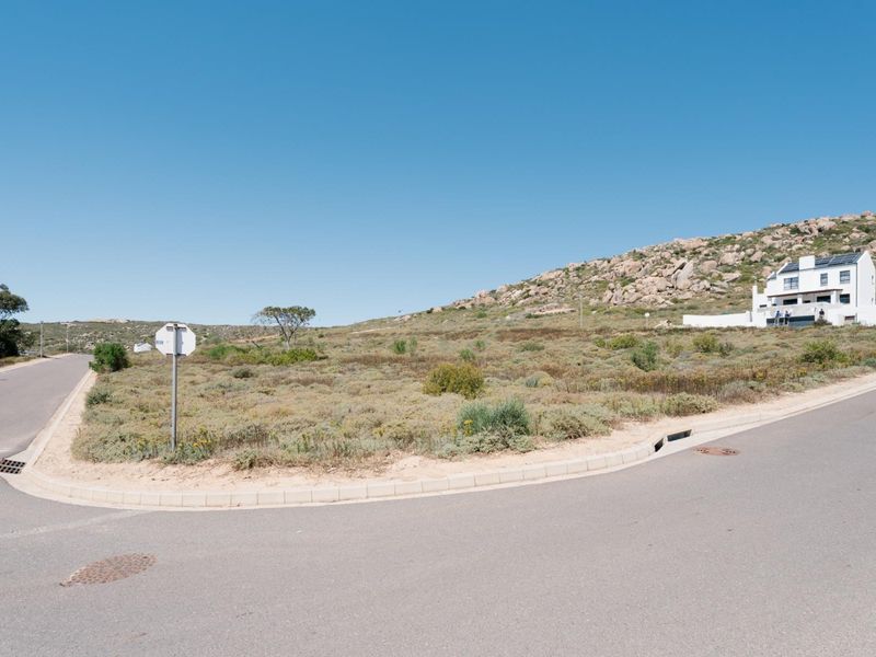 A street-level view of a vacant residential corner plot featuring established road infrastructure, curbing, and stormwater drainage in a coastal landscape setting.