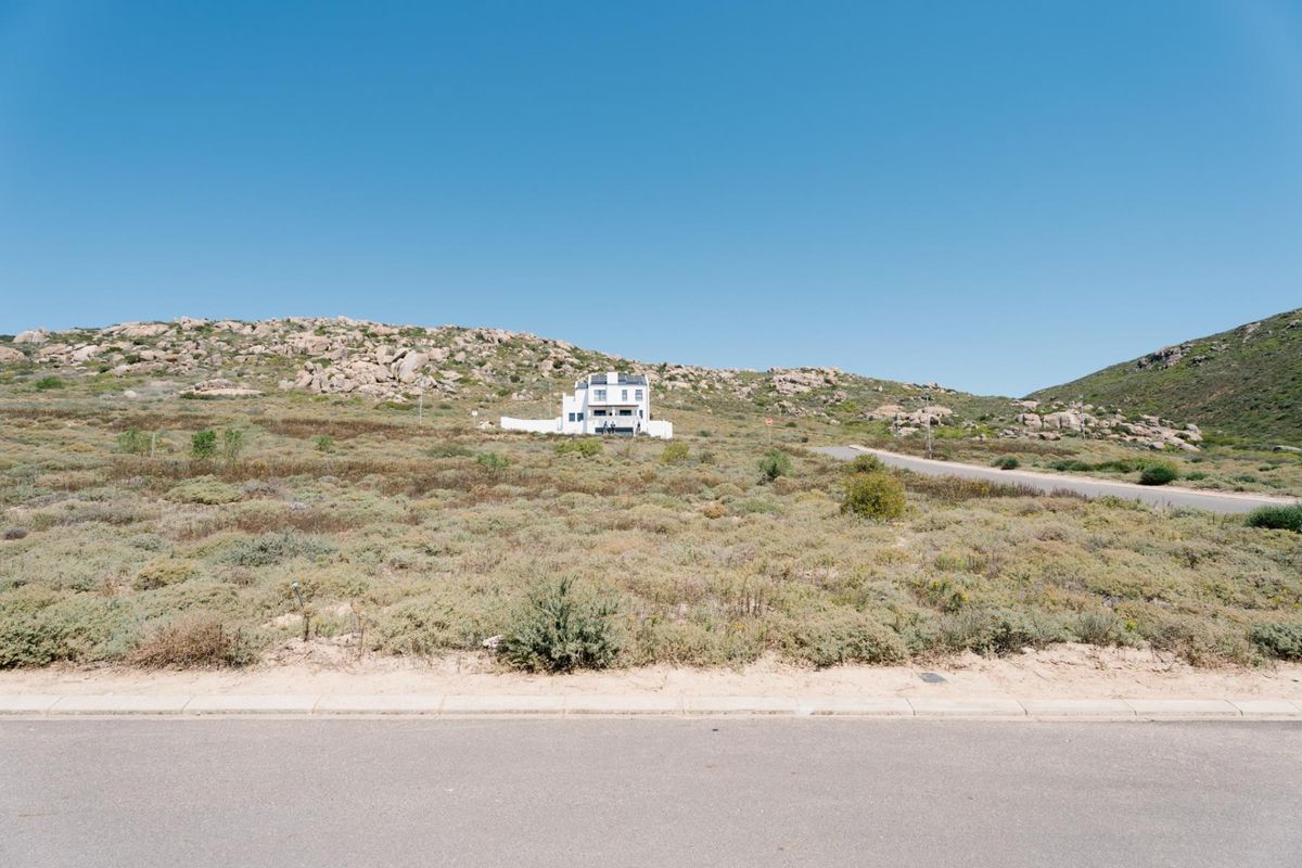 A wide-angle view of a vacant residential plot featuring natural coastal vegetation, bordering a paved road with a distant house visible in the background.