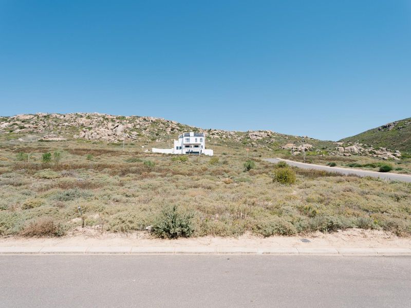 A wide-angle view of a vacant residential plot featuring natural coastal vegetation, bordering a paved road with a distant house visible in the background.