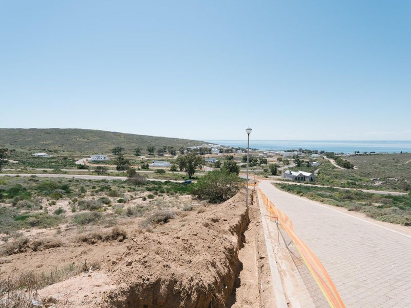Elevated view overlooking a coastal residential area with a paved road, street lighting, and ongoing infrastructure work in the foreground.
