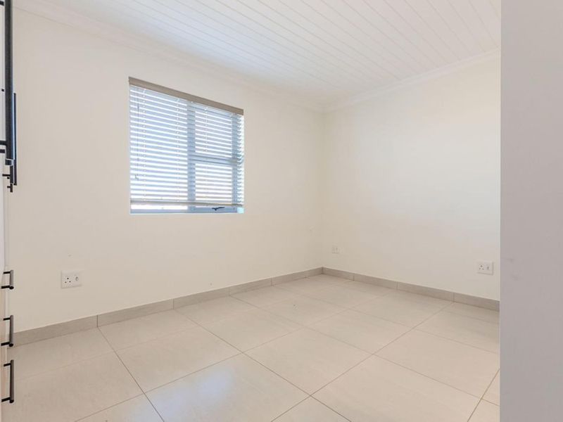 A bright, empty bedroom featuring neutral tiled flooring, a tongue and groove ceiling, and a window fitted with venetian blinds.