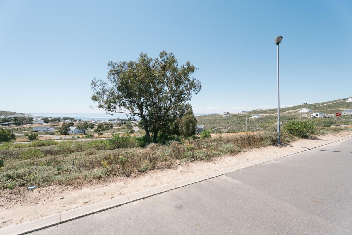 A street-level view of a vacant residential plot featuring a paved road, concrete curbing, and a street light, with surrounding coastal landscape views.