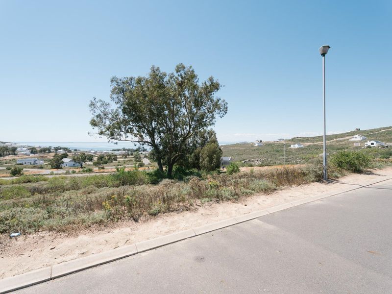 A street-level view of a vacant residential plot featuring a paved road, concrete curbing, and a street light, with surrounding coastal landscape views.