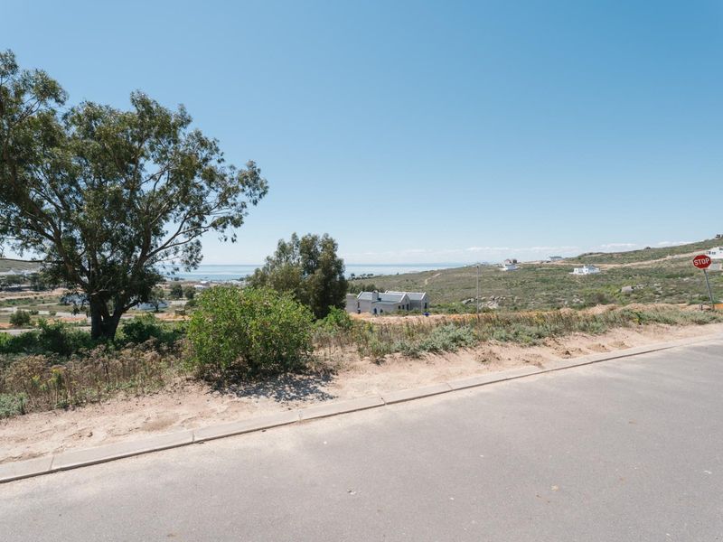 A street-level view of a vacant residential plot featuring natural coastal vegetation and distant ocean views.