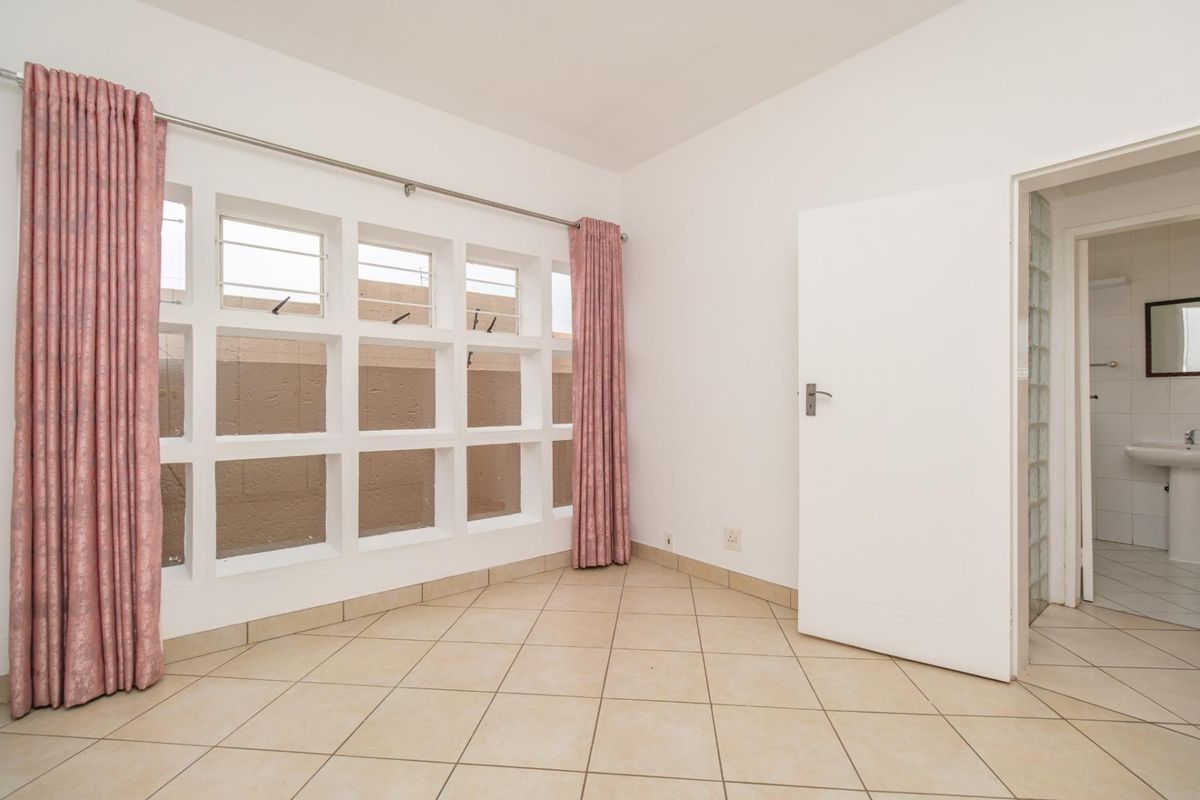 A bright, empty bedroom featuring tiled flooring, a large grid-style window, and direct access to an en-suite bathroom with a glass block feature wall.