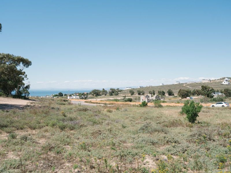 A scenic view overlooking a coastal landscape with natural vegetation, distant houses, and a glimpse of the ocean under a clear blue sky.