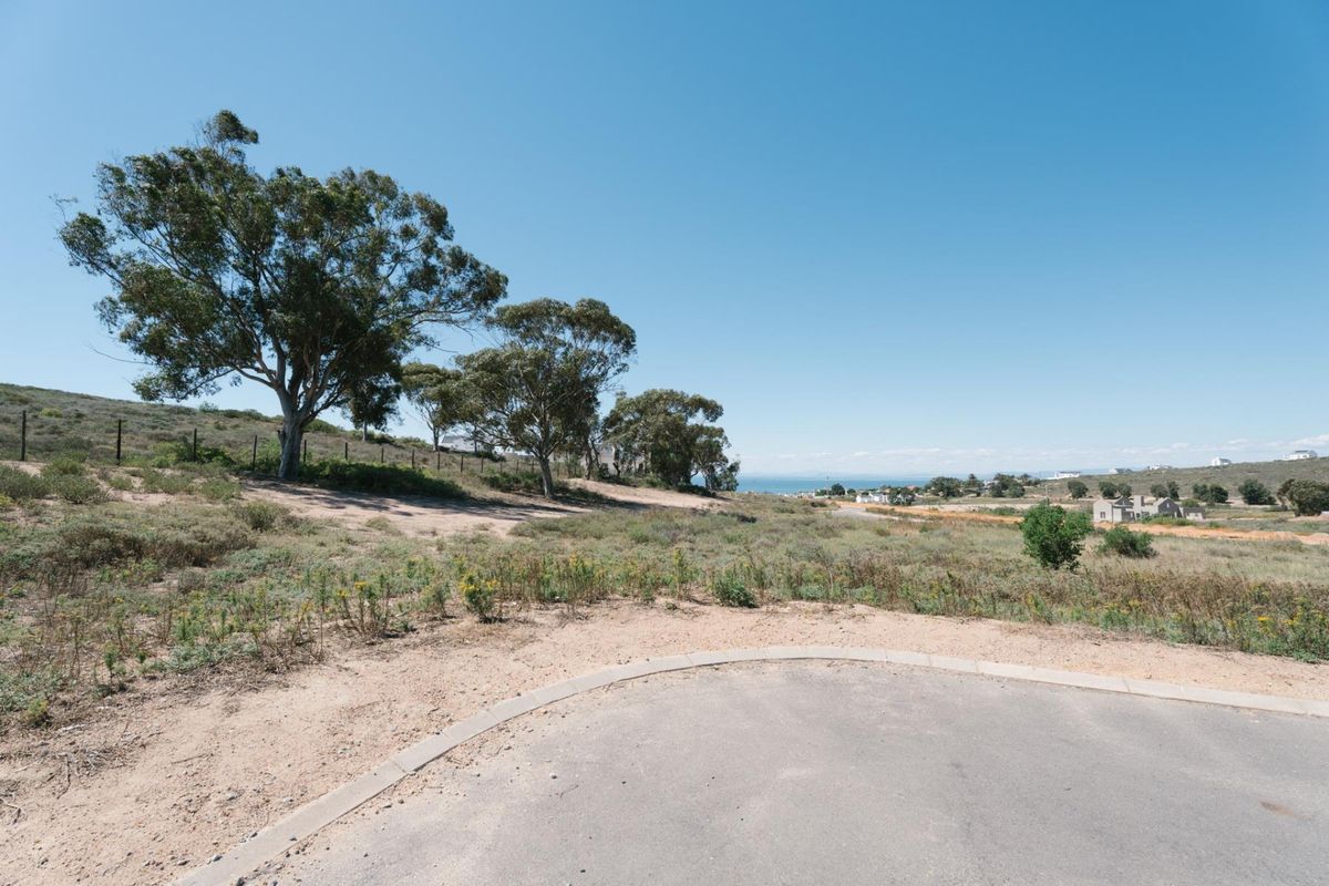 A vacant residential plot featuring natural vegetation, mature trees, and distant coastal views, accessible from a paved road.