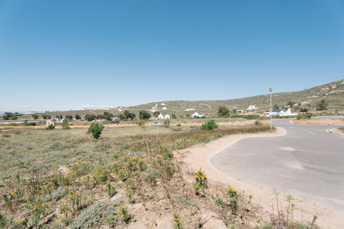 A wide-angle view of a vacant residential plot adjacent to a paved road, featuring natural landscape and surrounding hillside scenery.