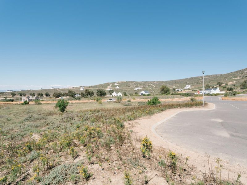 A wide-angle view of a vacant residential plot adjacent to a paved road, featuring natural landscape and surrounding hillside scenery.
