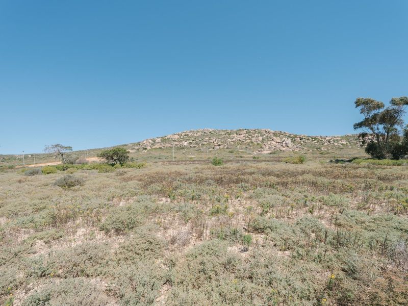 A wide-angle view of the vacant residential land featuring natural coastal vegetation and a rocky hillside backdrop under a clear blue sky.