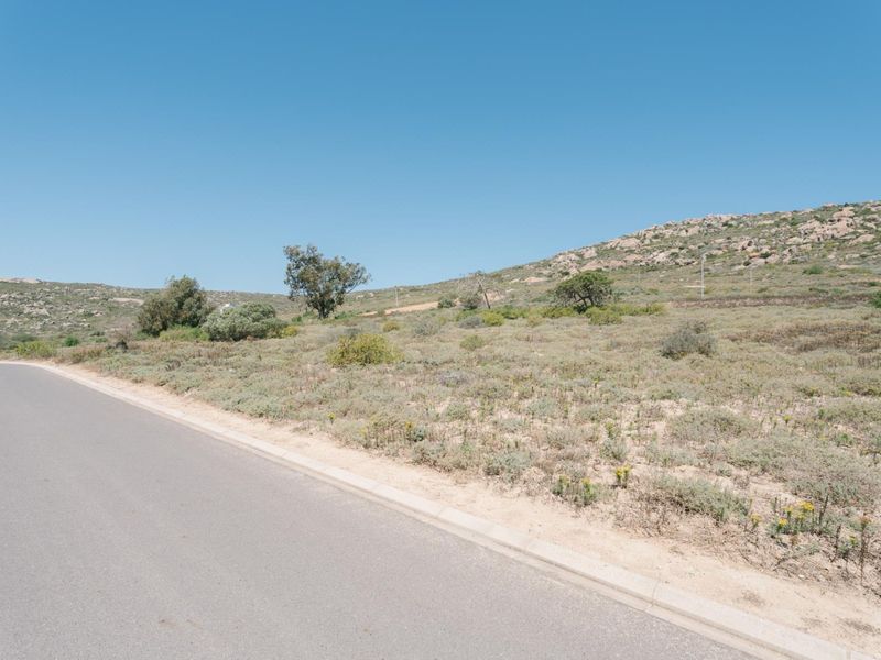 A view of the vacant residential land from the adjacent asphalt road, featuring natural vegetation and a rocky hillside backdrop.