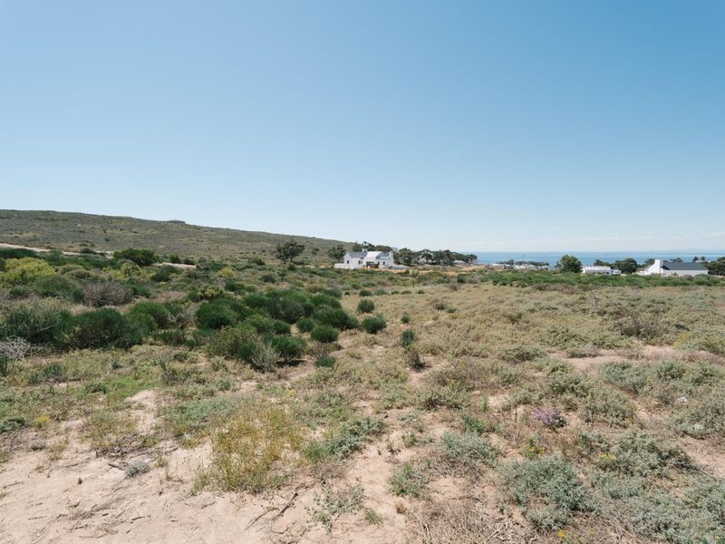 A wide-angle view of a vacant residential plot featuring natural coastal vegetation with distant ocean views and surrounding residential properties.