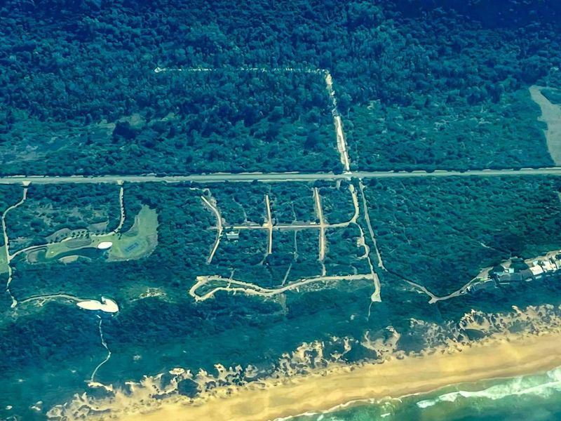 Aerial view of a residential land development site featuring cleared plots, road infrastructure, and proximity to the coastline and forest.